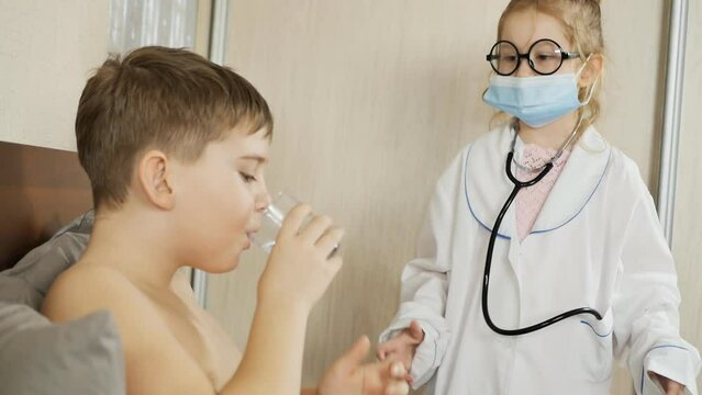 school age brother and sister play hospital. The boy swallowed a pill and washed it down with water, the girl in a dressing gown, mask, glasses and with a stethoscope takes care of him. slow motion