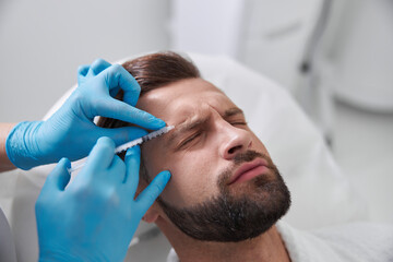Ppatient grimaces of pain while careful cosmetologist does injection of lifting filler in his brow in clinic