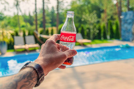 Man hand with a tattoo holds a glass bottle with coca-cola on the background of the pool and trees on a sunny day