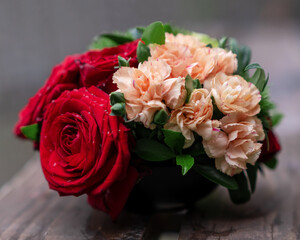A small bouquet of red roses, delicate pink carnations and green leaves, on a wooden brown background.