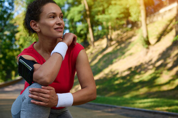 Beautiful Hispanic woman, athlete in sportswear relaxing during jog in the city park, smiles looking aside, enjoying morning workout on summer sunny day