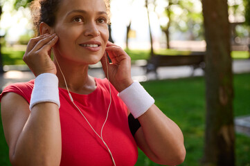 Attractive middle aged active African American woman, sportswoman putting on earphones, wearing...