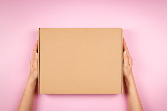 Female hands holding brown cardboard box on pastel pink background. Top view to mockup parcel box. Packaging, shopping, delivery concept