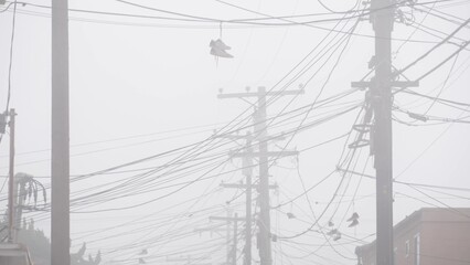 Sneakers canvas shoes hanging on power line, fog on city street, misty foggy weather in California,...