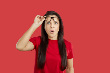 Beautiful young brunette woman impressed by something extraordinary looks at camera with surprised, shocked, astonished face expression, lifts her glasses and says WOW. Studio shot on red background - obrazy, fototapety, plakaty