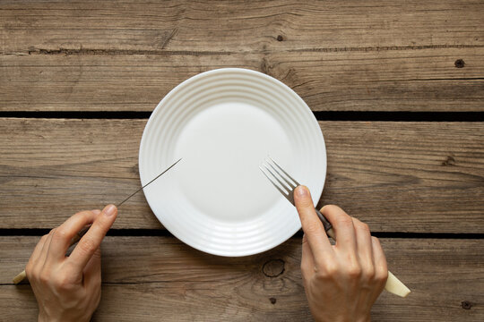 One white plate and fork with a knife in hands on an old wooden table in the kitchen at home, restaurant business