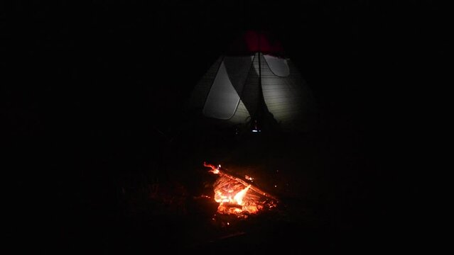 camp fire is burning in the night and people in the tent, light on inside, crackling wooden in fire, they are talking about sleeping in turkish