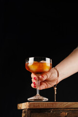 Female hand reaching for Scotch Whiskey or rum with ice cubes in cocktail glass with stem on wooden board on black background