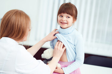 Fototapeta premium Cheerful female doctor checking patient little girl by stethoscope in hospital