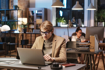 Young Caucasian man in eyeglasses and jacket sitting at desk and using laptop while working In Dark Office