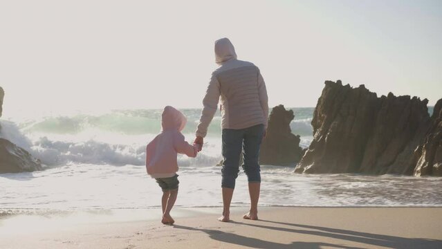 Mother and child playing ocean beach at spring, dressed warm clothes barefoot handheld video effect. Family with one child traveling at sea shore Mom and daughter spending time together 