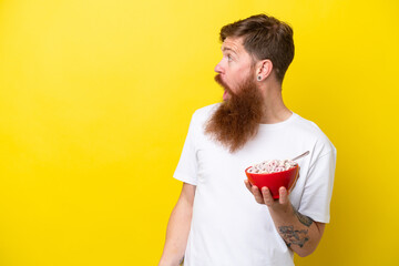Redhead man with beard eating a bowl of cereals isolated on yellow background with surprise facial...