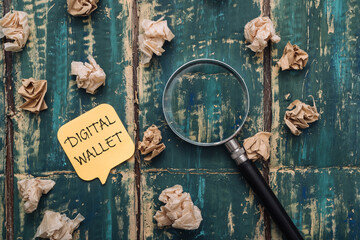 DIGITAL WALLET text on a sticky note on a green wooden background, crumpled notes and a magnifying glass lie nearby