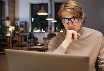 Serious thoughtful young manager in glasses working in dark office and using laptop while turning report in by deadline