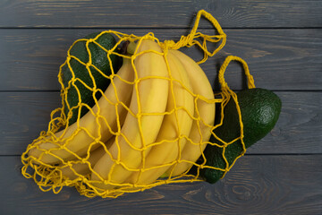 A yellow knitted string bag with bananas and avocado lies on a gray wooden background. The concept of zero waste, replacement of plastic bags.