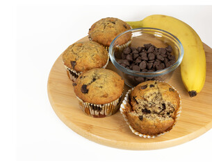 Banana chocolate chip muffins homemade a a partial round wooden board with a ripe banana and a glass dish of chocolate chips against a white background.