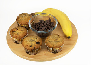 Banana chocolate chip muffins on a round wooden board with the delicious banana and chocolate chip ingredients displayed on a white background.