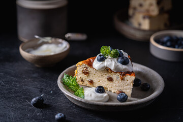 Baked homemade cottage cheese casserole or pudding with raisins serving with blueberry and sour cream on dark background. Close-up.
