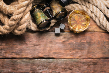 Whiskey in the drinking glass, binoculars and mooring rope on the brown wooden flat lay table...