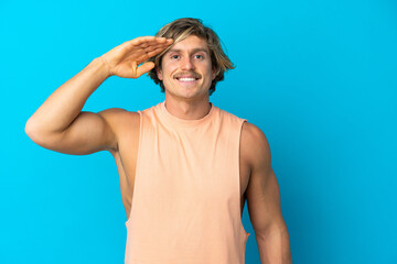 Handsome blonde man isolated on blue background saluting with hand with happy expression