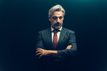 Portrait of attractive senior businessman wearing suit standing under the light over dark background.