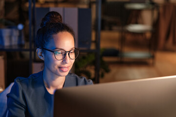 Positive young multiethnic woman in glasses using modern computer while working late in office