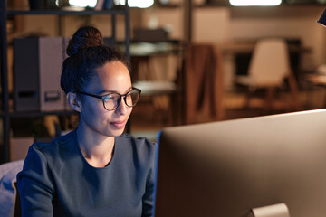 Obraz premium Content young biracial female office employee in glasses using computer while working at night