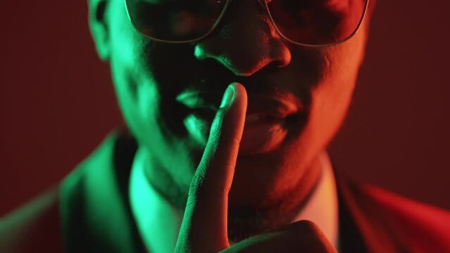 Shush gesture. Man secret. Keep quiet. Closeup of unrecognizable gentleman showing shh with finger on lips in green neon light isolated on dark red.