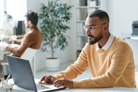 Serious Young Mixed Race Businessman In Yellow Sweater Working With Website On Laptop In Modern Office