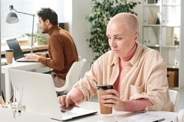 Content young Caucasian businesswoman in casual outfit drinking coffee and using laptop while working online