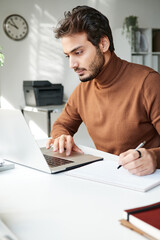 Concentrated young middle eastern man with stubble sitting at table and using laptop while drawing sketch in office