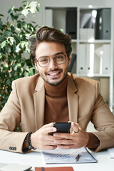 Portrait of cheerful young middle eastern manager in eyeglasses and bracelets sitting with smartphone at desk in office
