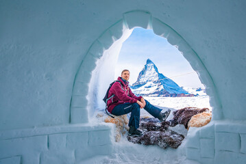 Portrait of young tourist resting at entrance of igloo. Famous snowcapped matterhorn peak is in background. Man enjoying winter in alpine region.