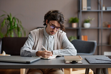 Obraz premium Horizontal medium portrait of young adult man wearing comfortable casual outfit and eyeglasses sitting at desk writing something in notebook