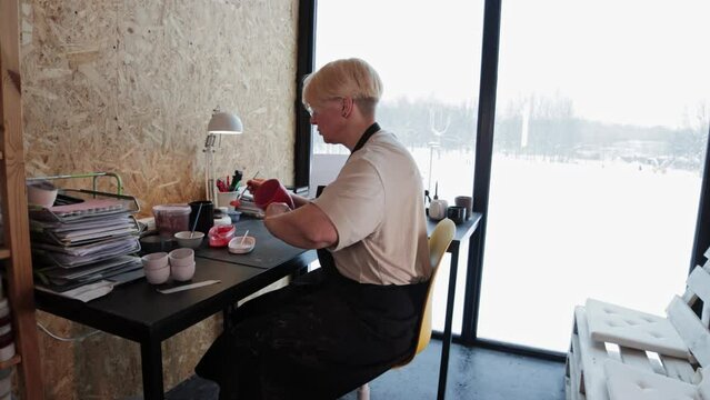 An elderly woman painting ceramic mug with red color