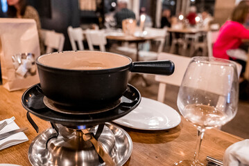Fototapeta na wymiar Heated pot of cheese fondue on burner. Wineglass and plates arranged on table. Gourmet swiss fondue dinner at luxurious ski resort during winter.