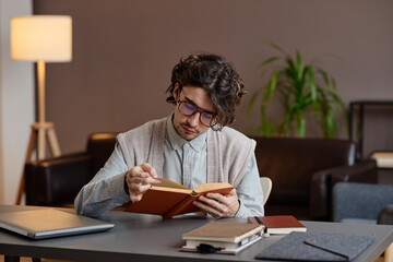 Horizontal medium shot of modern professional psychologist sitting at desk in his office reading book while waiting for client