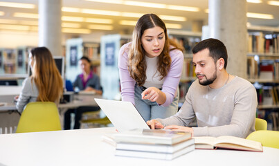 Two friendly young adult male and female students preparing for exam together in university library, using textbooks and laptop