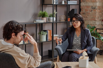 Horizontal medium shot of professional psychotherapist offering her client glass of water to calm...