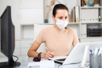 Woman in protective mask sitting at workplace with computer in her office