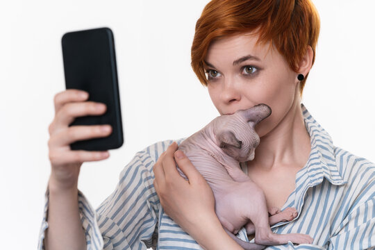 Woman holding and kissing kitten, taking selfie self portrait photos on smartphone. Young adult redhead hipster dressed striped white-blue shirt. Part of series. Studio shot on white background.