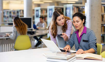 Caring woman helping friend to prepare for exams in public library