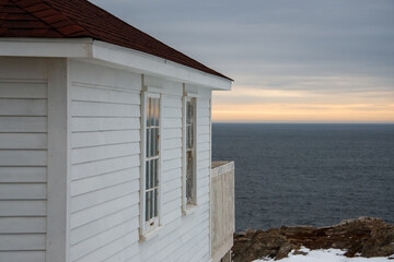 The exterior of a white wooden clapboard building with two glass windows, a black roof and a wooden...