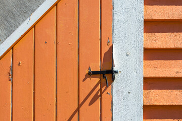 A vintage black metal latch on an orange wooden door made of narrow boards. There's thick white trim around the door and a small window. The exterior wall is made of wooden clapboard siding. 