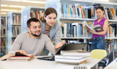 Young positive interested couple of students spending time together in library, reading books and browsing educational websites on laptop