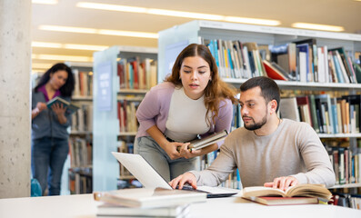 Positive man and woman spending time together in library, talking, work on laptop
