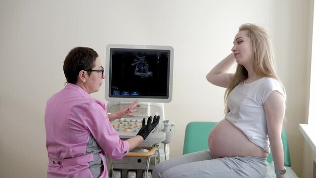 A female doctor examines a pregnant girl in the hospital. The gynecologist listens to the pregnant girl and records the results of the tests to the pregnant patient in the ultrasound room