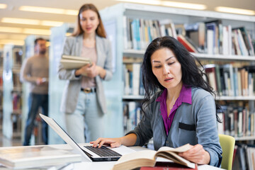 Obraz premium Motivated young adult asian female student studying in university library, using books and laptop while preparing for course ..