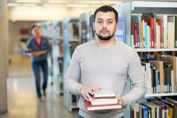 Portrait of confident successful young adult bearded student holding stack of books, standing in...