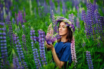 Fototapeta premium A young brunette woman in a flower wreath made of lupine with a bouquet of lupines smiles gently and looks at the camera in a flower meadow, inhales the fragrance of flowers.